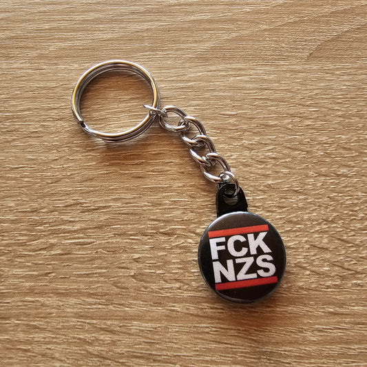 „Schlüsselanhänger mit rundem Anhänger und der Aufschrift ‚FCK NZS‘ in weißer Blockschrift auf schwarzem Hintergrund.“
