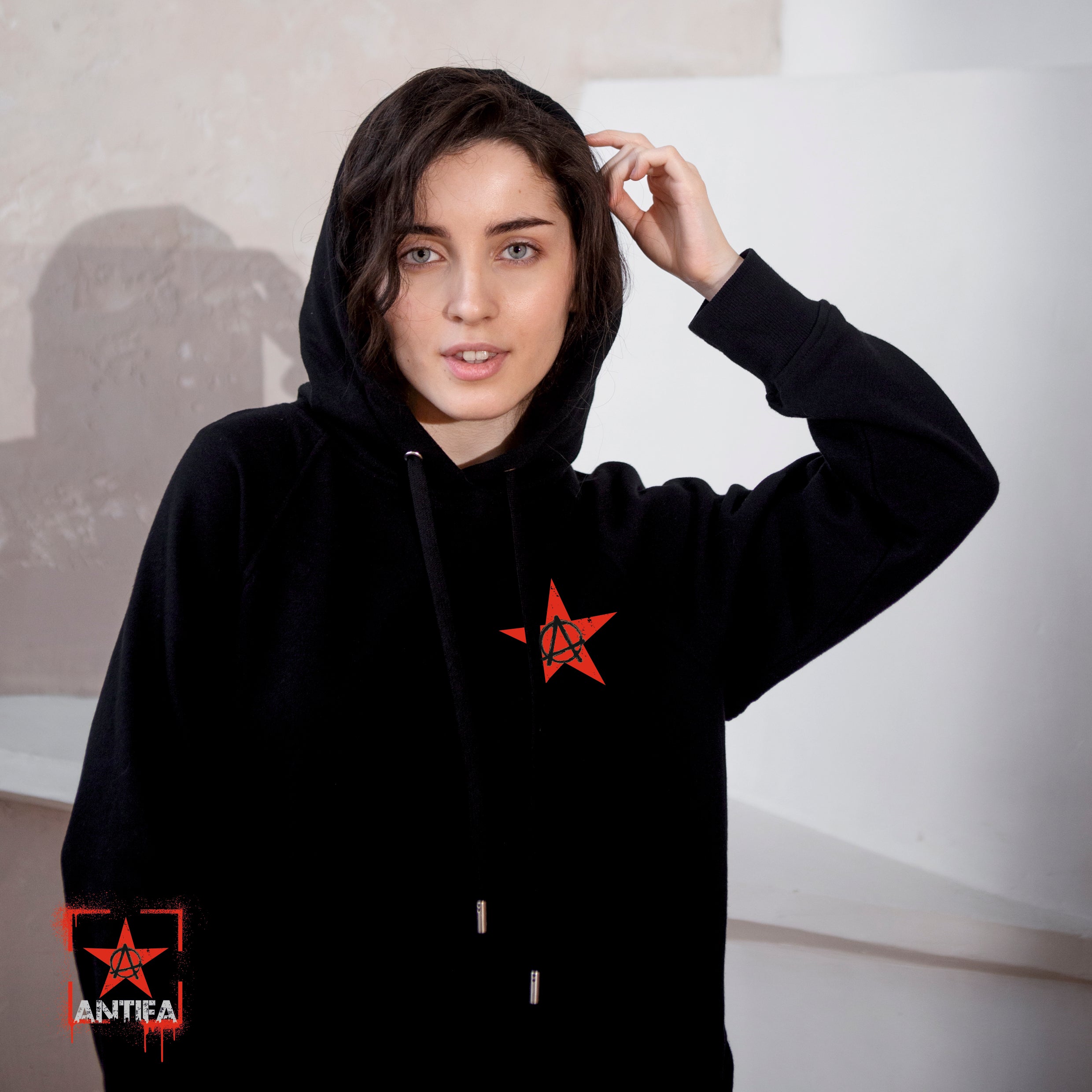 Person trägt schwarzen Hoodie mit rotem Stern und eingekreistem „A“-Symbol auf der Brust – ein Zeichen für anarchistische und antifaschistische Haltung. In der Ecke erscheint zusätzlich das Wort „ANTIFA“ in Weiß neben dem Symbol.