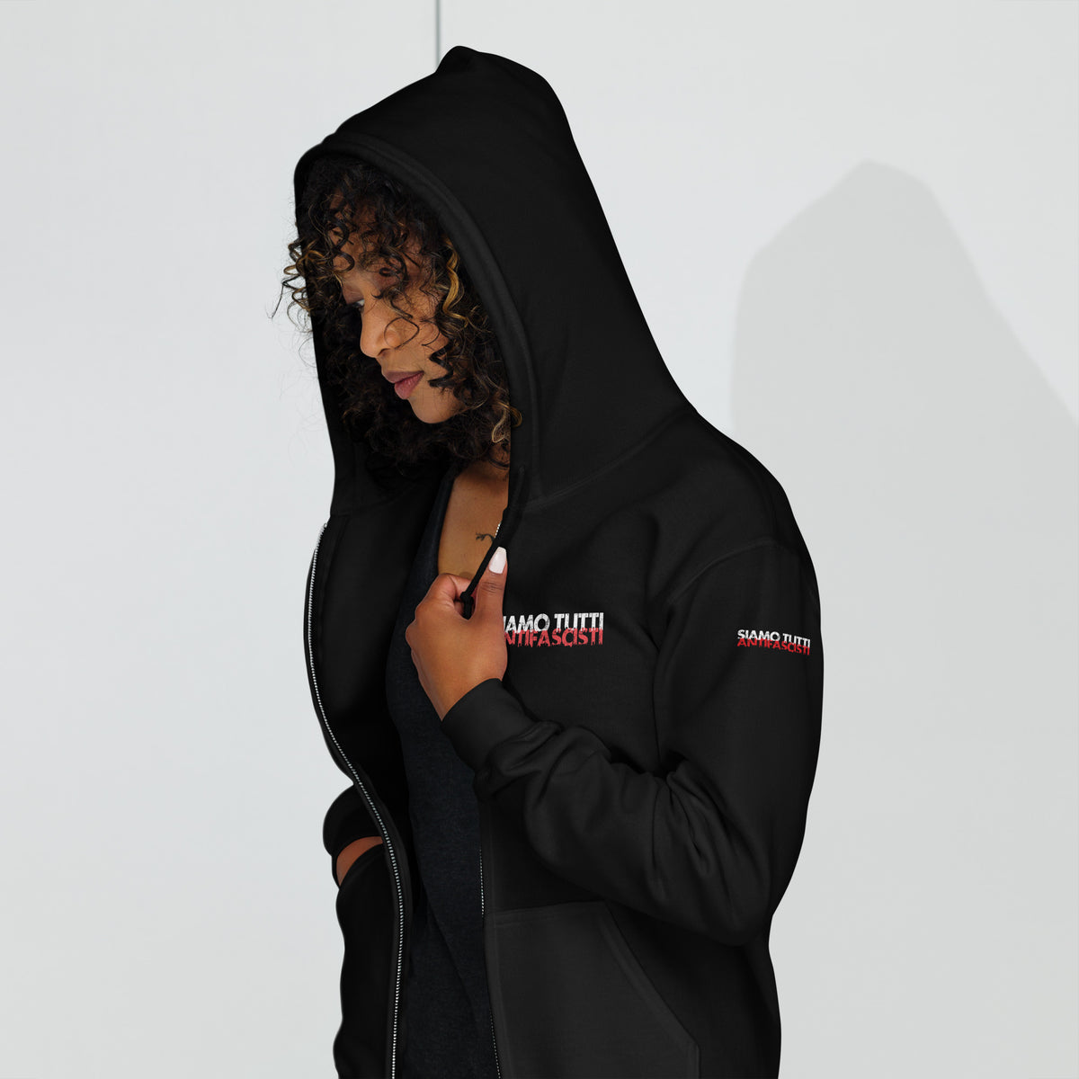 „Person trägt schwarzen Zip‑Hoodie mit Kapuze und Fronttasche, Aufdruck ‚SIAMO TUTTI ANTIFASCISTI‘ in Weiß und Rot auf Brust und Ärmel – antifaschistisches Statement in lebendiger Tragesituation.“